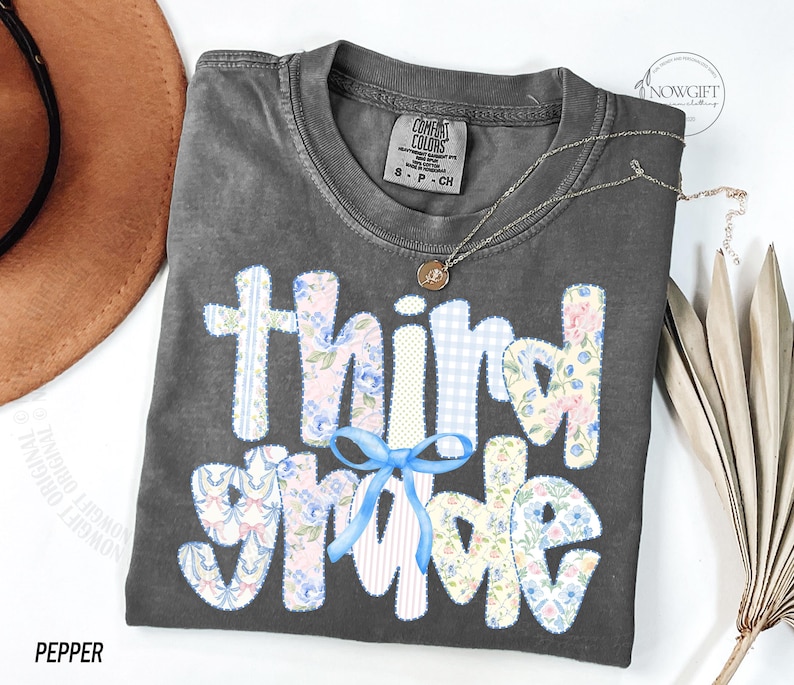 Puede incluir: Una camiseta gris Comfort Colors con las palabras "Third Grade" en un dise&ntilde;o de patchwork. Las letras est&aacute;n en patrones florales, de cuadros y a rayas, con un lazo azul. Tambi&eacute;n se ven un collar dorado y un sombrero marr&oacute;n.