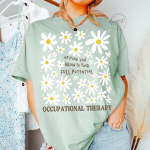 May include: A light green t-shirt with a daisy design and the text "Helping You Grow To Your Full Potential" and "Occupational Therapy" in brown. The shirt has short sleeves and a relaxed fit.