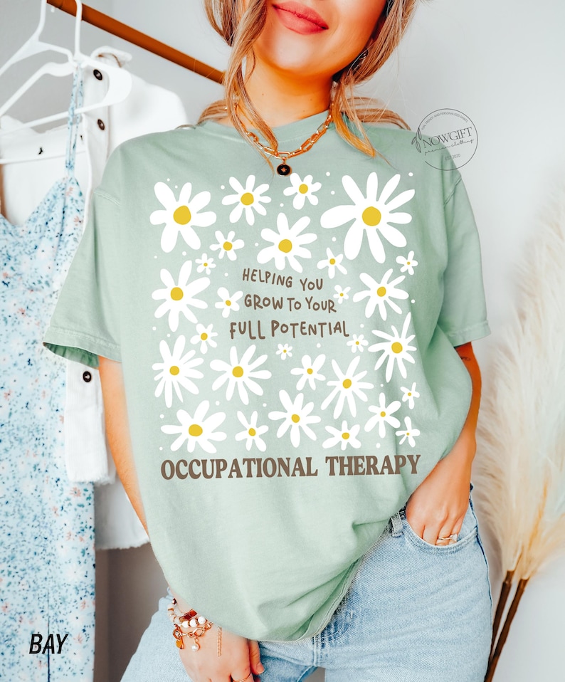 May include: A light green t-shirt with a daisy design and the text "Helping You Grow To Your Full Potential" and "Occupational Therapy" in brown. The shirt has short sleeves and a relaxed fit.