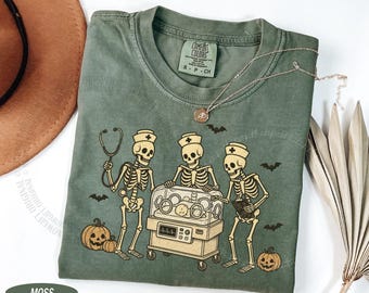 Op de afbeelding: Een mosgroen T-shirt met korte mouwen en een afbeelding van drie skeletten verkleed als medisch personeel rondom een couveuse met een baby erin. Het ontwerp bevat twee pompoenen, diverse kleine vleermuizen en een stethoscoop.