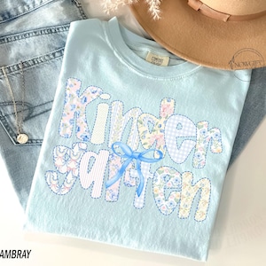 Pu&ograve; includere: Una maglietta azzurra con la scritta "Kindergarten" in un design patchwork. Le lettere sono realizzate con motivi floreali, a righe e a quadretti, con un fiocco blu. La maglietta &egrave; piegata sopra dei jeans blu.