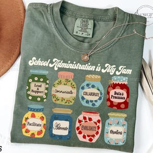 May include: A green t-shirt with the text "School Administration is My Jam" printed on it. The text is surrounded by illustrations of jars with different words on them, such as "Lead & Support", "Communicate", "Collaborate", "Data & Processes", "Facilitate", "Advocate", "Evaluate", and "Mentors".