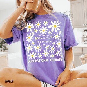 May include: Violet-colored t-shirt with a daisy design and the text "Helping You Grow To Your Full Potential. Occupational Therapy." The shirt has short sleeves and a relaxed fit. The word "VIOLET" is printed at the bottom left.
