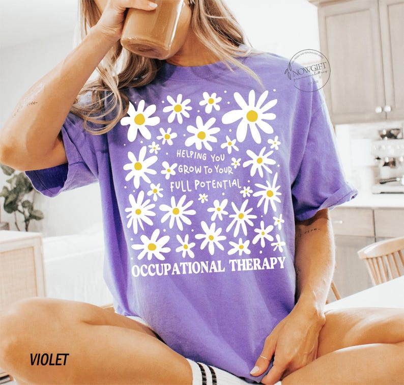 May include: Violet-colored t-shirt with a daisy design and the text "Helping You Grow To Your Full Potential. Occupational Therapy." The shirt has short sleeves and a relaxed fit. The word "VIOLET" is printed at the bottom left.