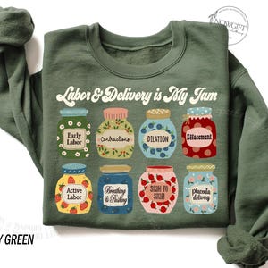 May include: A green sweatshirt with the text "Labor & Delivery is My Jam" in a retro font. The text is surrounded by nine illustrated jars of jam, each with a different label related to labor and delivery, such as "Early Labor", "Contractions", and "Effacement".