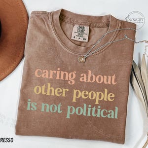 May include: A brown t-shirt with the text "caring about other people is not political" printed in rainbow colors.