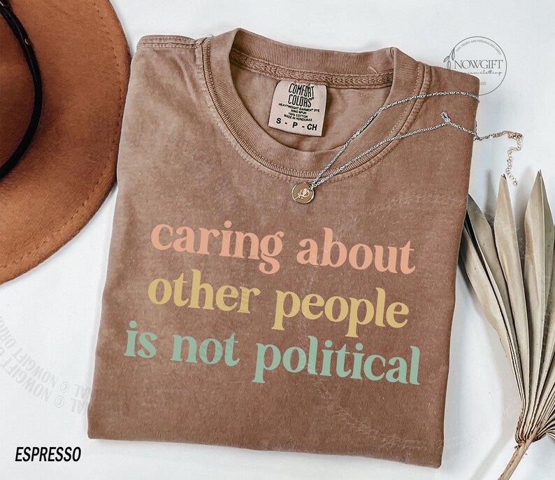 May include: A brown t-shirt with the text "caring about other people is not political" printed in rainbow colors.