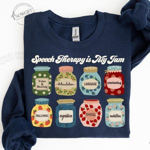 May include: Navy blue sweatshirt with the text "Speech Therapy is My Jam" in white. The sweatshirt features a row of nine colorful jars with different speech therapy related words on them.