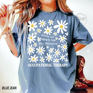 May include: Blue jean colored t-shirt with a daisy flower design. The shirt has the text "Helping You Grow To Your Full Potential" and "Occupational Therapy" in white. The shirt is a comfortable, casual style.