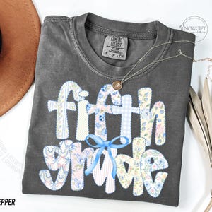 May include: A dark gray t-shirt with the words "fifth grade" in a patchwork design. The letters are decorated with floral and gingham patterns, and a blue bow is at the bottom. A gold necklace and a brown hat are also visible.