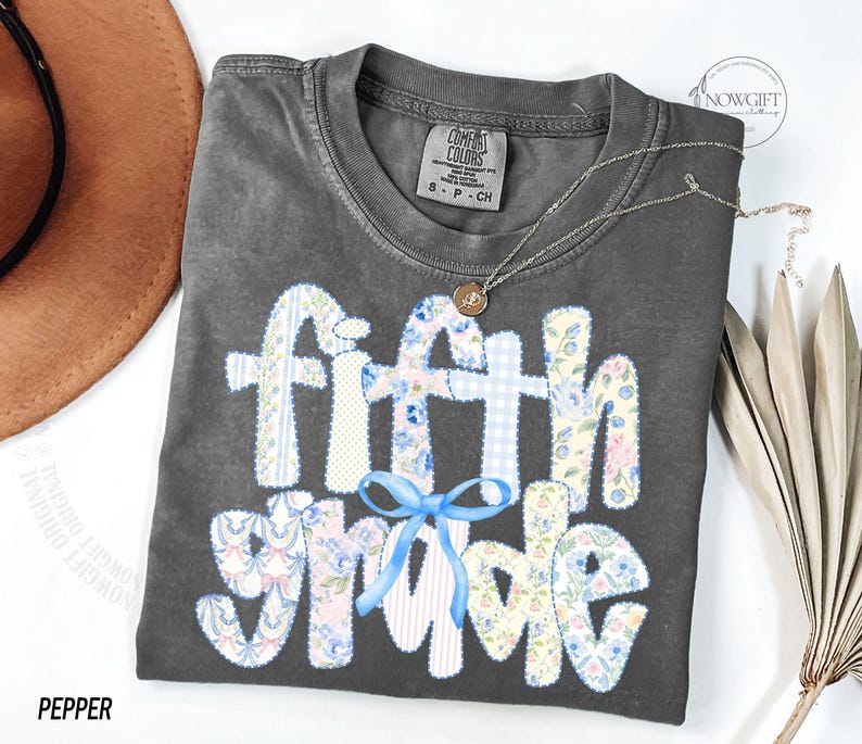 May include: A dark gray t-shirt with the words "fifth grade" in a patchwork design. The letters are decorated with floral and gingham patterns, and a blue bow is at the bottom. A gold necklace and a brown hat are also visible.