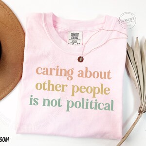May include: Pink t-shirt with the text "caring about other people is not political" in pastel colors.