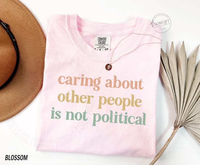 May include: Pink t-shirt with the text "caring about other people is not political" in pastel colors.