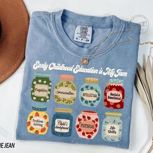Puede incluir: Una camiseta de mezclilla azul con el texto "Early Childhood Education is My Jam" e ilustraciones de frascos con diferentes aspectos del desarrollo de la primera infancia, como la cognici&oacute;n, la comunicaci&oacute;n, la inteligencia, lo social y lo emocional, la resoluci&oacute;n de problemas, el desarrollo del ni&ntilde;o, lo f&iacute;sico y las habilidades para la vida.