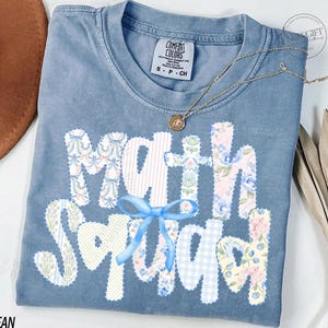 May include: A blue jean Comfort Colors t-shirt with the words "Math Squad" in a patchwork design. The letters are decorated with floral and patterned fabrics, and a blue bow is attached. A gold necklace with a pendant is draped across the shirt.