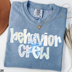 May include: A blue jean Comfort Colors t-shirt with the words "behavior crew" in a floral and gingham pattern. A light blue bow is centered under the word "behavior". A gold necklace with a pendant is draped on the shirt.