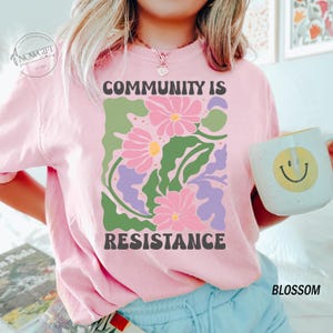 May include: Pink t-shirt with a floral design and the text "Community is Resistance".