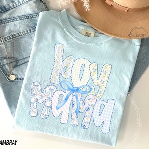 Puede incluir: Camiseta azul claro con las palabras "boy mama" en un dise&ntilde;o de patchwork. Las letras presentan patrones florales, de lunares y cuadros, con un lazo azul. La palabra "CHAMBRAY" est&aacute; impresa en la parte inferior.
