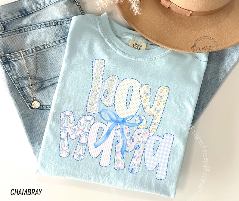 Puede incluir: Camiseta azul claro con las palabras "boy mama" en un dise&ntilde;o de patchwork. Las letras presentan patrones florales, de lunares y cuadros, con un lazo azul. La palabra "CHAMBRAY" est&aacute; impresa en la parte inferior.
