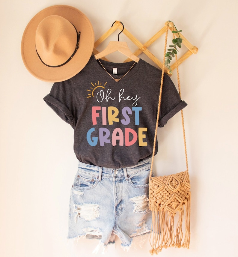 May include: A dark gray t-shirt with a yellow sun graphic and the text "Oh hey FIRST GRADE" in rainbow colors. The shirt is hanging on a wooden hanger with a straw hat and a woven bag.