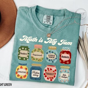 May include: Light green t-shirt with the text "Math is My Jam" printed on the front. The text is in a retro font and is surrounded by illustrations of jars with different math-related themes, such as "Numbers", "Measurement", "Algebraic Thinking", "Geometry", "Operations", "Problem Solving", "Probability", and "Data Analysis".