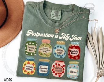 Może przedstawiać: Koszulka w kolorze mchu z napisem "Postpartum is My Jam". Koszulka przedstawia kolorowe ilustracje słoików z dżemem z etykietami takimi jak "Physical Care" i "Emotional Support". Widoczny jest złoty naszyjnik.