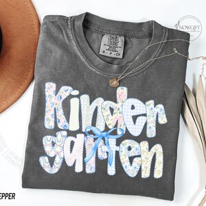 Pu&ograve; includere: Una maglietta Comfort Colors grigio scuro con la scritta "Kindergarten" in un design patchwork. Le lettere sono realizzate con tessuti floreali e a quadretti, con un fiocco blu. Una collana d'oro con un ciondolo &egrave; sulla maglietta.