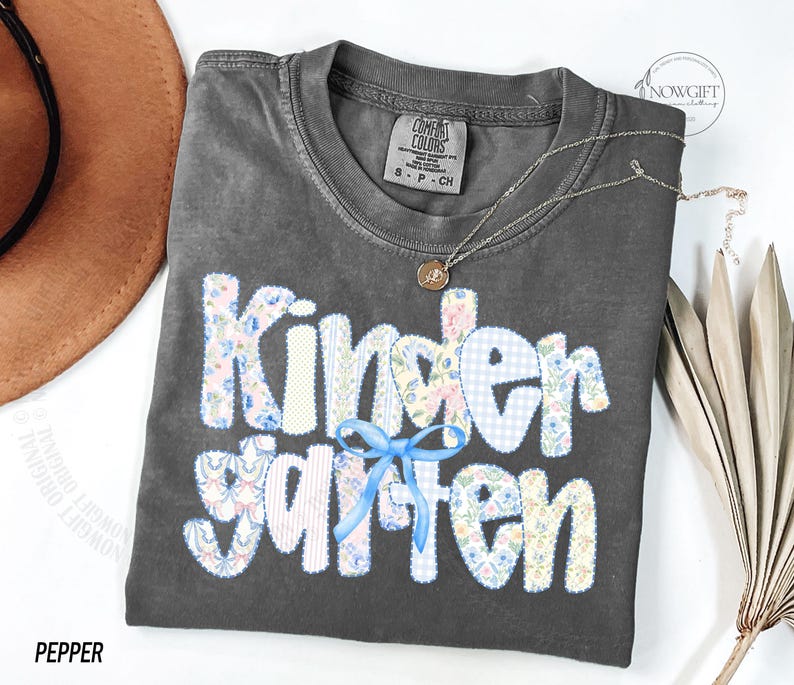 Pu&ograve; includere: Una maglietta Comfort Colors grigio scuro con la scritta "Kindergarten" in un design patchwork. Le lettere sono realizzate con tessuti floreali e a quadretti, con un fiocco blu. Una collana d'oro con un ciondolo &egrave; sulla maglietta.