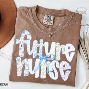 May include: Espresso-colored t-shirt with the words "Future Nurse" in a patchwork design. The letters are white with floral and gingham patterns. A light blue bow is beneath the word "Nurse". A silver necklace with a small pendant is on the shirt.