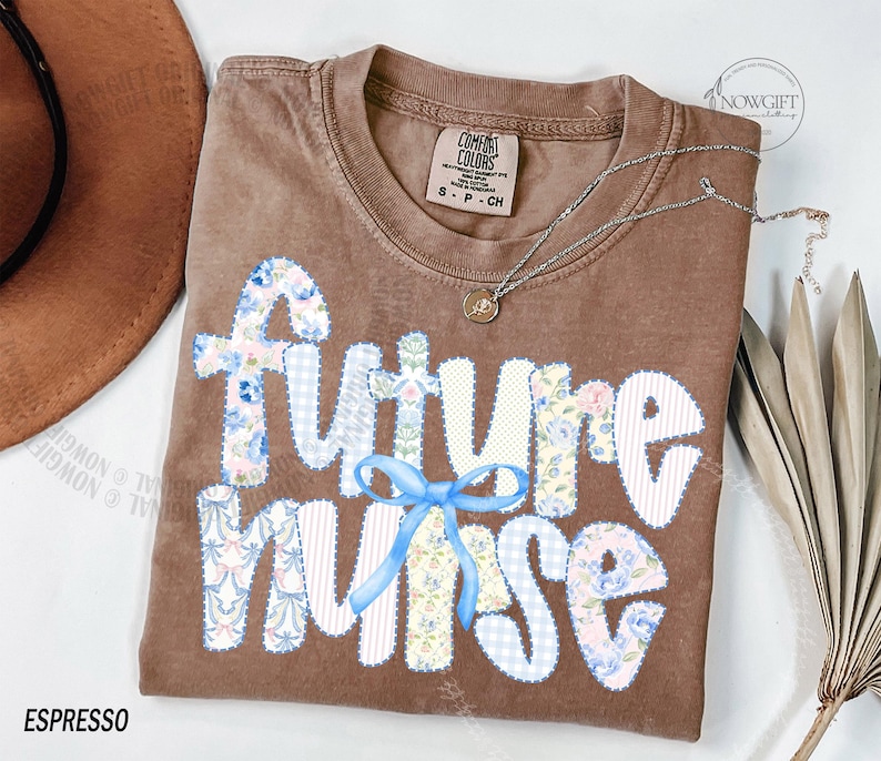 May include: Espresso-colored t-shirt with the words "Future Nurse" in a patchwork design. The letters are white with floral and gingham patterns. A light blue bow is beneath the word "Nurse". A silver necklace with a small pendant is on the shirt.