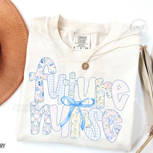 May include: Ivory-colored t-shirt with the words "Future Nurse" in a patchwork design. The letters are filled with floral and gingham patterns in blue, pink, and white, with a blue ribbon. A gold necklace with a pendant is also visible.