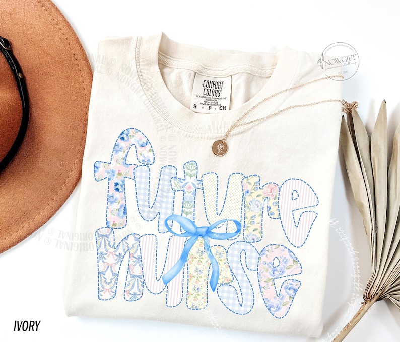 May include: Ivory-colored t-shirt with the words "Future Nurse" in a patchwork design. The letters are filled with floral and gingham patterns in blue, pink, and white, with a blue ribbon. A gold necklace with a pendant is also visible.