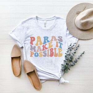 May include: A white t-shirt with the text "PARAS MAKE IT POSSIBLE" in rainbow colored letters. The shirt is laid flat on a white textured surface with a brown hat, two brown shoes, and a sprig of eucalyptus.