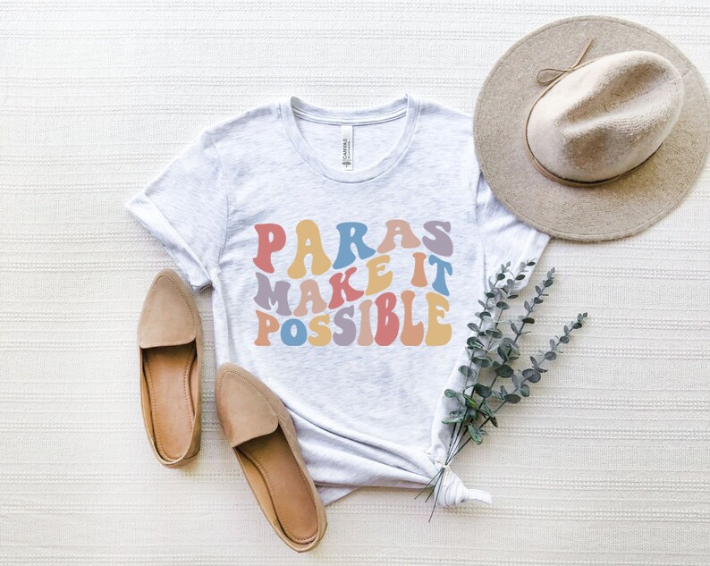 May include: A white t-shirt with the text "PARAS MAKE IT POSSIBLE" in rainbow colored letters. The shirt is laid flat on a white textured surface with a brown hat, two brown shoes, and a sprig of eucalyptus.