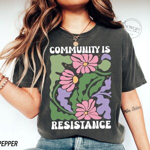 May include: A dark grey t-shirt with a graphic print of pink and purple flowers and green leaves. The text "COMMUNITY IS RESISTANCE" is printed in white on the shirt.