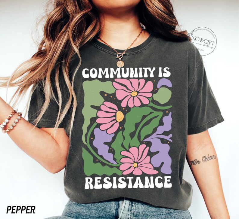 May include: A dark grey t-shirt with a graphic print of pink and purple flowers and green leaves. The text "COMMUNITY IS RESISTANCE" is printed in white on the shirt.
