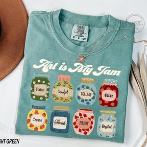 Op de afbeelding: Een lichtgroen T-shirt met de tekst "Art is My Jam" in een retro-lettertype. Het shirt heeft een ontwerp van acht kleurrijke potten met verschillende kunstgerelateerde woorden erop, zoals "Verf", "Beeldhouwen", "Collage", "Kleur", "Cre&euml;ren", "Schetsen", "Drukken" en "Digitaal".