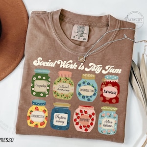 May include: A brown t-shirt with the text "Social Work is My Jam" in a retro font. The shirt features nine jars with different designs and text, including "Empathy", "Cultural Awareness", "Organization", "Advocacy", "Communication", "Problem Solving", "Support", and "Active Listener".