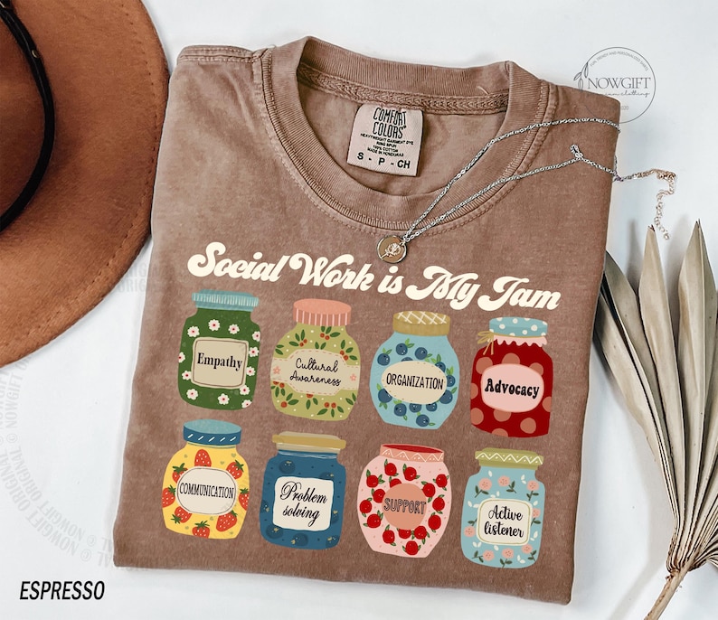 May include: A brown t-shirt with the text "Social Work is My Jam" in a retro font. The shirt features nine jars with different designs and text, including "Empathy", "Cultural Awareness", "Organization", "Advocacy", "Communication", "Problem Solving", "Support", and "Active Listener".