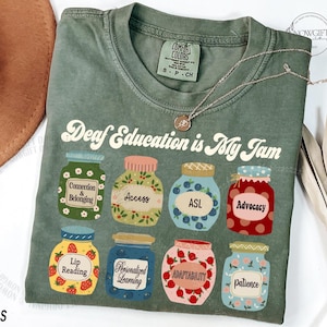 May include: Moss green t-shirt with the text "Deaf Education is My Jam" and colorful jar illustrations. Each jar has a different word inside, including "Connection & Belonging", "Access", and "ASL". A gold necklace is draped on the shirt.