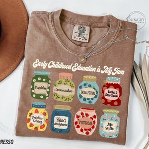 Puede incluir: Una camiseta marr&oacute;n con el texto "Early Childhood Education is My Jam" impreso en ella. El texto est&aacute; rodeado de ilustraciones de tarros con diferentes etiquetas, incluyendo "Cognitive", "Communication", "Intellectual", "Social & Emotional", "Problem-Solving", "Child's Development", "Physical", y "Life Skills".