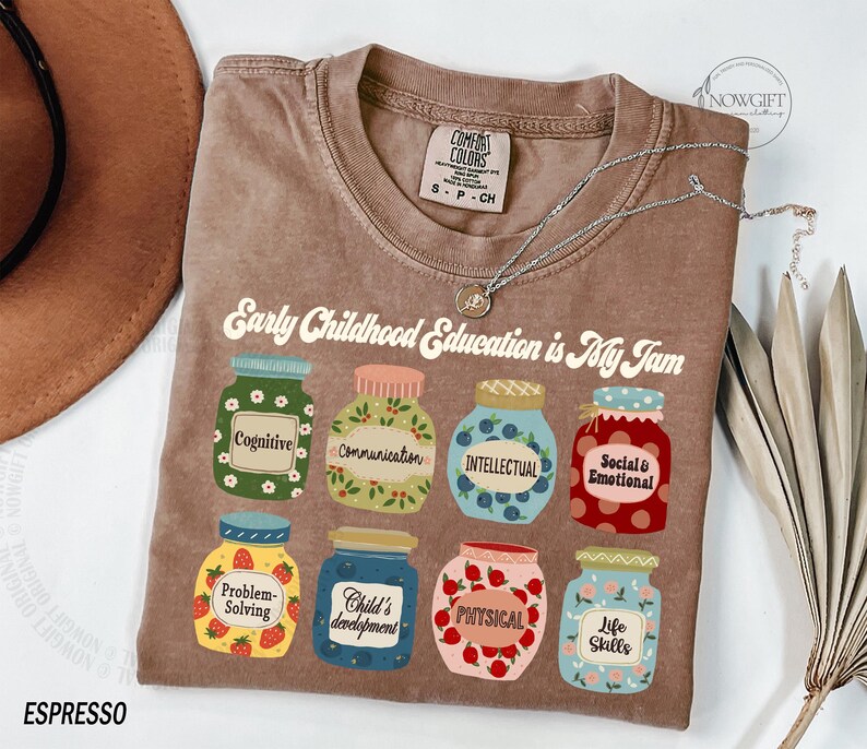 Puede incluir: Una camiseta marr&oacute;n con el texto "Early Childhood Education is My Jam" impreso en ella. El texto est&aacute; rodeado de ilustraciones de tarros con diferentes etiquetas, incluyendo "Cognitive", "Communication", "Intellectual", "Social & Emotional", "Problem-Solving", "Child's Development", "Physical", y "Life Skills".