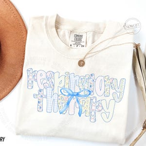Pu&ograve; includere: T-shirt color avorio con la scritta "respiratory therapy" in un design patchwork con motivi floreali e gingham. Un fiocco azzurro &egrave; centrato sulla maglietta. Una collana d'oro con un ciondolo &egrave; drappeggiata sulla maglietta.