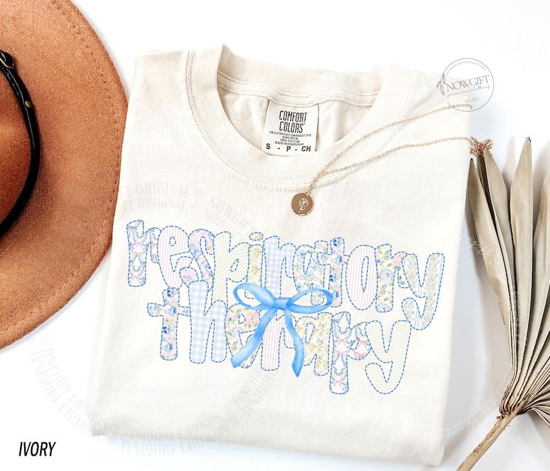 Pu&ograve; includere: T-shirt color avorio con la scritta "respiratory therapy" in un design patchwork con motivi floreali e gingham. Un fiocco azzurro &egrave; centrato sulla maglietta. Una collana d'oro con un ciondolo &egrave; drappeggiata sulla maglietta.