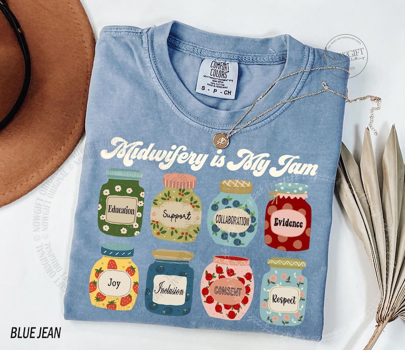 May include: A light blue Comfort Colors t-shirt with the phrase "Midwifery is My Jam" and jar graphics. Each jar has a different word: Education, Support, Collaboration, Evidence, Joy, Inclusion, Consent, and Respect. A gold necklace is draped across the shirt.