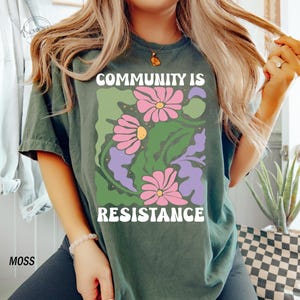 May include: A dark green t-shirt with a floral design and the text "Community is Resistance" in white letters.