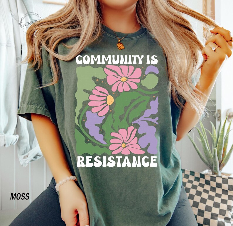 May include: A dark green t-shirt with a floral design and the text "Community is Resistance" in white letters.