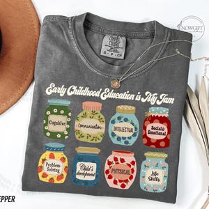 Puede incluir: Una camiseta gris con el texto "Early Childhood Education is My Jam" e ilustraciones de tarros con diferentes aspectos de la educaci&oacute;n de la primera infancia, como la cognici&oacute;n, la comunicaci&oacute;n, la intelectual, la social y la emocional, la resoluci&oacute;n de problemas, el desarrollo del ni&ntilde;o, la f&iacute;sica y las habilidades para la vida.