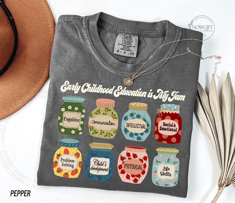 Puede incluir: Una camiseta gris con el texto "Early Childhood Education is My Jam" e ilustraciones de tarros con diferentes aspectos de la educaci&oacute;n de la primera infancia, como la cognici&oacute;n, la comunicaci&oacute;n, la intelectual, la social y la emocional, la resoluci&oacute;n de problemas, el desarrollo del ni&ntilde;o, la f&iacute;sica y las habilidades para la vida.