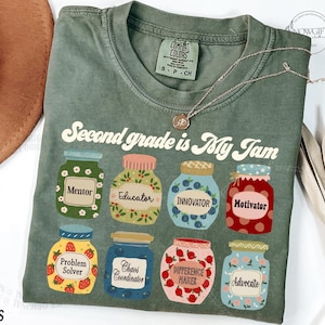 May include: A green t-shirt with the text "Second grade is my jam" and illustrations of jars with different labels, including "Mentor", "Educator", "Innovator", "Motivator", "Problem Solver", "Chaos Coordinator", "Difference Maker", and "Advocate".